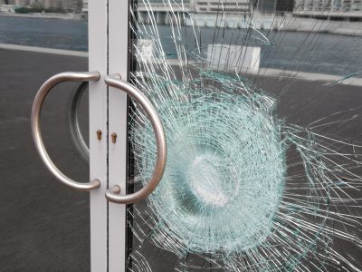 Broken window on business glass door shattered by vandalism Broken glass door panel with smashed area. Shattered window on business entrance of an office building facade cracked and damaged by vandalism. The doubled glass pane is silvered on the back reflecting the cracks and sidewalk.