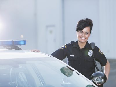 Female police officer standing next to patrol car A policewoman standing next to her patrol car, smiling and looking at the camera with her hands on her hips. She is wearing the standard navy blue policeuniform.